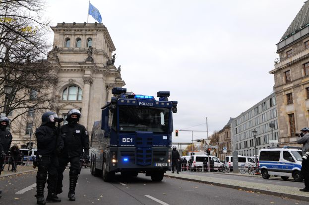 Eine Gruppe von Polizisten in Helmen und bewaffnet, die vor einem Gebäude mit Fenstern, Säulen, Bögen und einer Flagge stehen, mit Fahrzeugen auf der Straße, Bäumen, Laternenpfählen und einem bewölkten Himmel im Hintergrund.