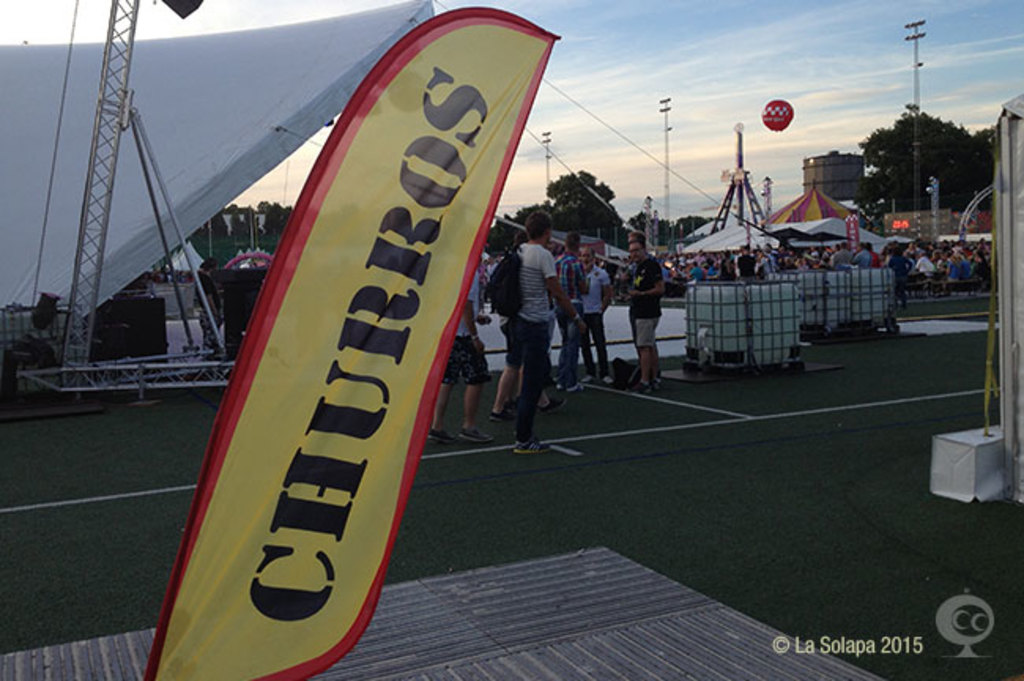 Ein Karnevalsbild mit Zelten, einem zentralen Mast, Menschen, Bäumen, Strommasten, Straßenbahnen auf der Straße und einem Eisenständer auf der linken Seite vor einem Himmel als Hintergrund.