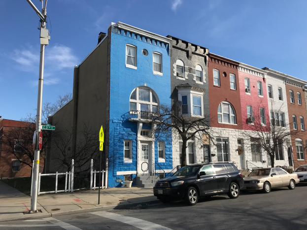 Eine Reihe bunter Häuser an einer Straßenkreuzung in Baltimore, Maryland, mit parkenden Autos, Bäumen, Pfählen, Brettern und einem Zaun, der die Straße säumt.
