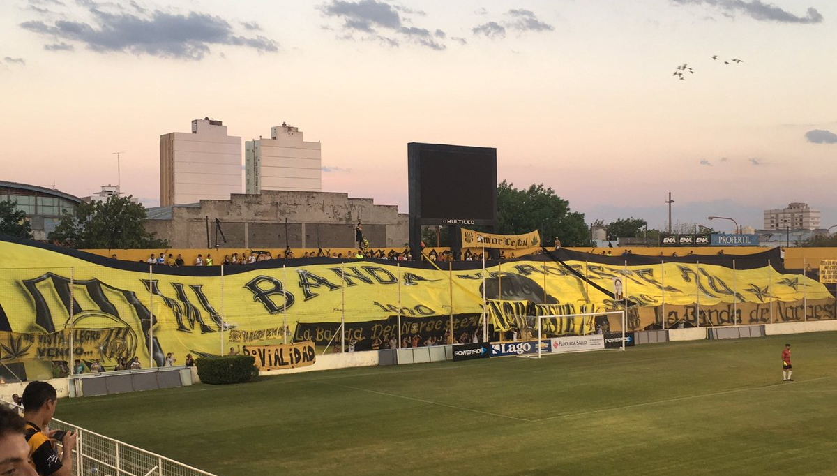 Ein Fußballfeld mit einer Gruppe von Menschen darauf, umgeben von einem Metallzaun, einem Torpfosten, einer Banner mit der Aufschrift "Boca Juniors vs Real Sociedad", einigen Pflanzen, einem Anzeigebildschirm, Pfählen, Baumrinde, Bäumen, Gebäuden und einem bewölkten Himmel.
