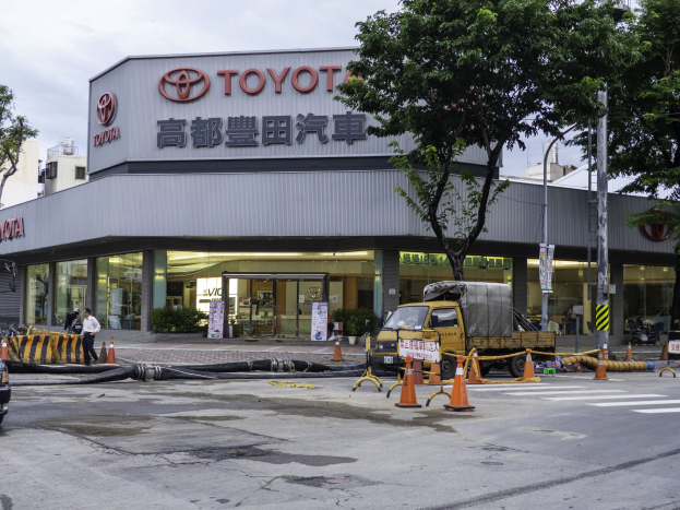Außenansicht eines Toyota-Autohauses in Tokio, Japan, mit Fahrzeugen auf der Straße, Verkehrskegeln, Rohren, Fußgängern, Bäumen, Strommasten, Beschilderung und umliegenden Gebäuden unter einem bewölkten Himmel.
