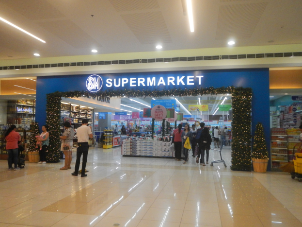 Eine Gruppe von Menschen steht vor einem Supermarkt, einige halten Tüten, mit einem Einkaufswagen, Topfpflanze, Regalpräsentationen, Schildern und Deckenleuchten im Hintergrund.