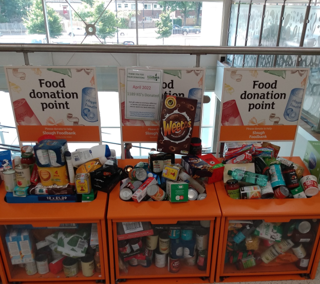 Orange Schübe mit Lebensmittelspenden gefüllt, darunter Flaschen, Dosen und Kisten, mit informativen Schildern im Hintergrund; Glasfenster zeigen Fahrzeuge, Bäume, Gebäude und Himmel.