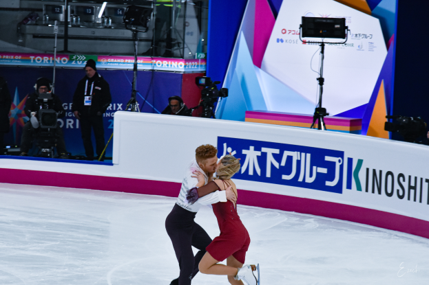 Ein Paar umarmt sich beim Eislaufen auf einer Eisbahn, mit einigen Menschen im Hintergrund, einige halten Kameras und eine Tafel mit Schrift darauf.