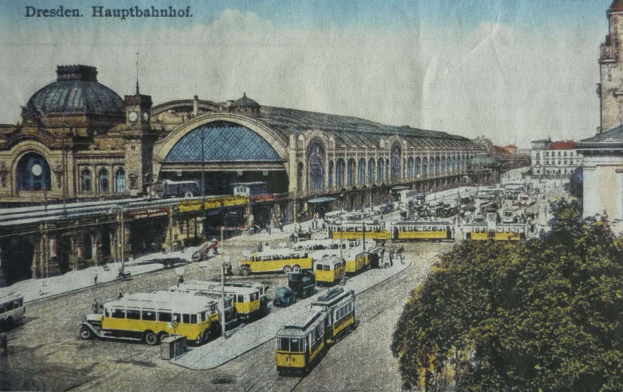 Ein sepiafarbenes Postkartenbild vom Dresdner Hauptbahnhof, das Pferdewagen, Fußgänger, Bäume, Gebäude und eine bewölkte Himmel zeigt.