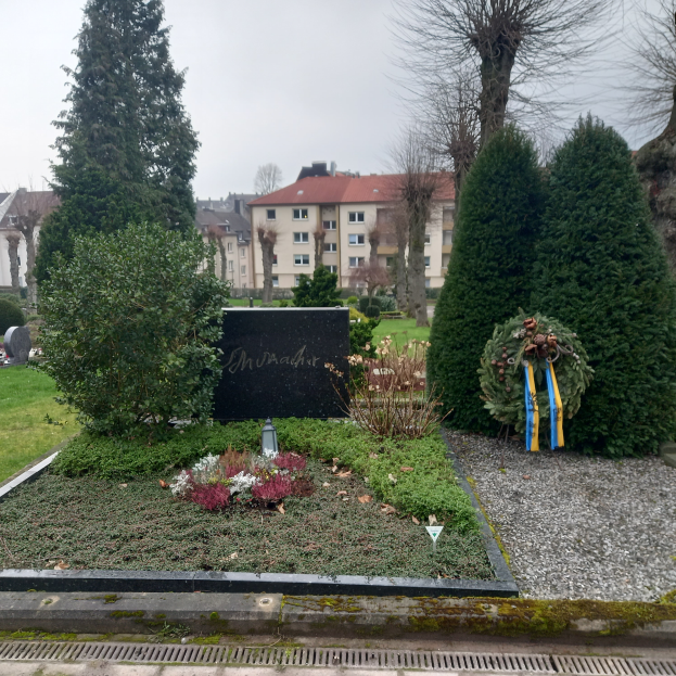 Ein Denkmal für Opfer des Zweiten Weltkriegs steht in einem Park, geschmückt mit einem Kränzchen und umgeben von Grün, mit Gebäuden im Hintergrund.