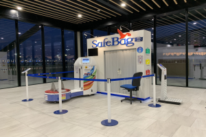 Sicherheitskontrolle für Gepäck am Flughafen mit Stuhl, Absperrgittern, Maschinen, Bildschirm, bedruckter Tafel, Glaswänden, Deckenleuchten und sichtbarem Boden.