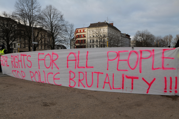 Eine Gruppe von Menschen hält ein Transparent mit der Aufschrift 'Rechte für alle Menschen Stoppt Polizeigewalt' auf dem Boden stehend mit einem Straßenschild, einem Schild, Bäumen, Gebäuden mit Fenstern und einem bewölkten Himmel im Hintergrund.