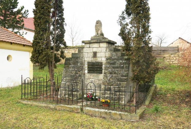 Ein steinernes Denkmal auf einer Rasenfläche, umgeben von einem Metallzaun und geschmückt mit Blumensträußen, mit Bäumen, Häusern und einer Wand im Hintergrund unter einem klaren blauen Himmel, als Holocaust-Gedenkstätte in Sibiu, Rumänien.