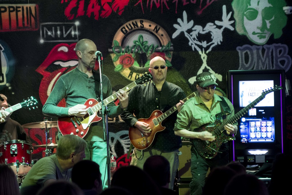 Drei Männer auf einer Bühne, die Gitarren spielen, mit einer Menge unten und einer gemalten Wand im Hintergrund.