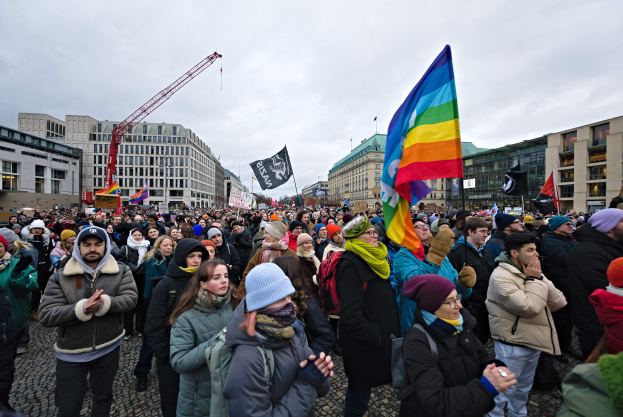 Eine große Gruppe von Menschen auf einer LGBTQ+-Rechtsdemo in Berlin, die Fahnen und Plakate schwenken, mit Gebäuden und einem Kran im Hintergrund unter einem bewölkten Himmel.