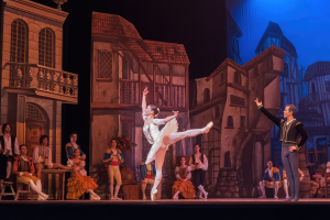 Zwei Tänzer performen auf einer Bühne im Royal Opera House in London, mit einem Publikum im Vordergrund und einem Bildschirm mit Gebäudeabbildungen im Hintergrund.