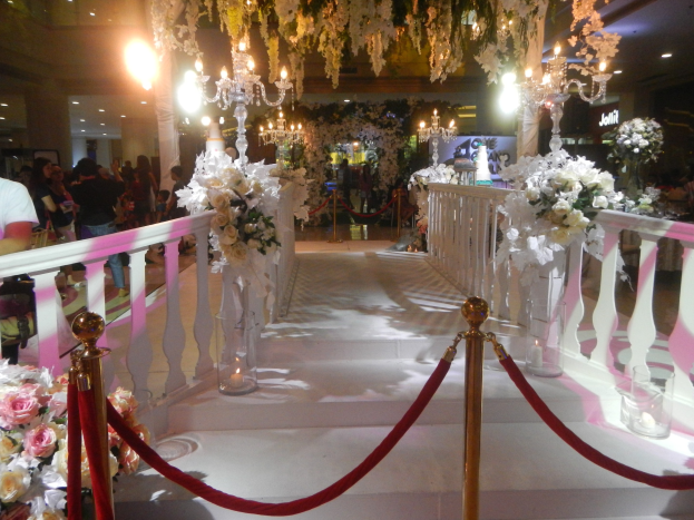 Eine Hochzeitszeremonie in einem Einkaufszentrum mit weißem Teppich, verzierten Geländern, Blumenschmuck und Deckenbeleuchtung, bei der zahlreiche Gäste anwesend sind.