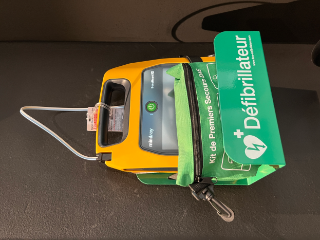 Ein gelb-grüner Defibrillator auf einem schwarzen Tisch mit einer grünen Tasche daneben, die Text enthält.