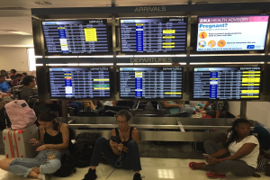 Menschen sitzen auf einem Flughafenvorfeld, einige halten Handys, mit verstreuten Taschen und Bildschirmen, die Zika-Virus-Informationen anzeigen, im Hintergrund.