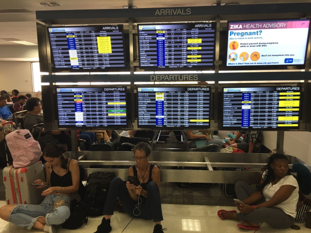 Menschen sitzen auf einem Flughafenvorfeld, einige halten Handys, mit verstreuten Taschen und Bildschirmen, die Zika-Virus-Informationen anzeigen, im Hintergrund.