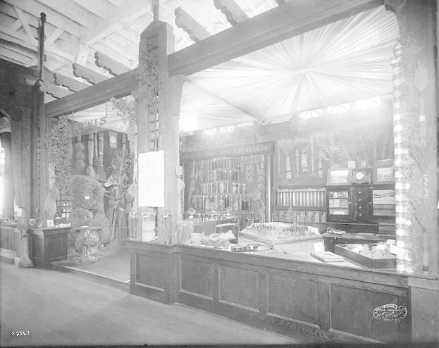 Ein altes Schwarz-Weiß-Foto einer Apotheke mit einem Tresen, der verschiedene Gegenstände und Regale mit Flaschen und anderen Gegenständen im Hintergrund zeigt.