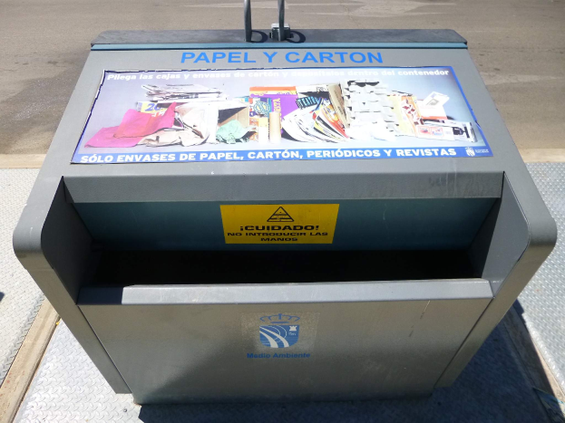 Ein Metallmüllcontainer am Straßenrand mit einem "Papel y Carton"-Plakat, Fahrzeuge im Hintergrund.