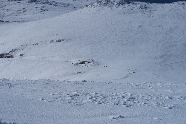 Ein schneebedeckter Berg mit ein paar Skifahrern, die hinunterfahren, unberührter Schnee und ein strahlend blauer Himmel, mit schneebedeckten Bergen im Hintergrund.