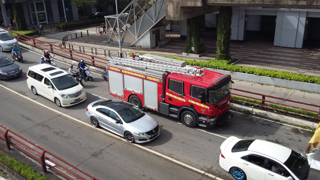 Feuerwehrfahrzeug fährt auf einer Straße mit Autos, Motorrädern und anderen Fahrzeugen, neben Geländern und Gebäuden mit Bäumen und Pfählen im Hintergrund.