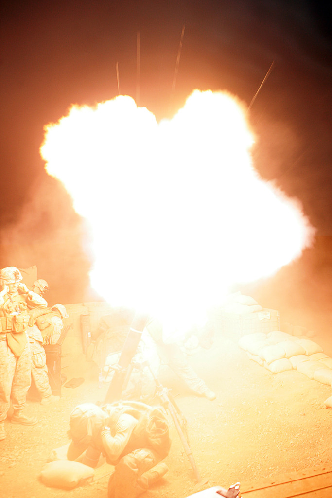 Mehrere Personen in Uniformen und Helmen auf einem Gelände mit einigen Taschen drumherum, mit einem Feuer in der Mitte der Szene.