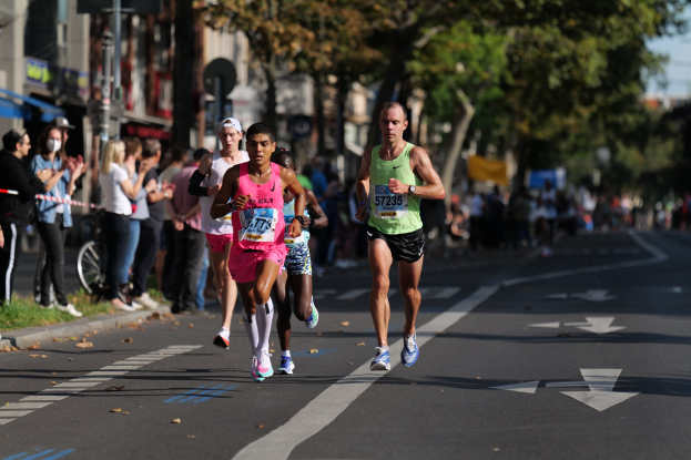 Gruppe von Läufern bei einem Marathon auf einer städtischen Straße mit Zuschauern, Grünflächen und urbanen Elementen im Hintergrund.