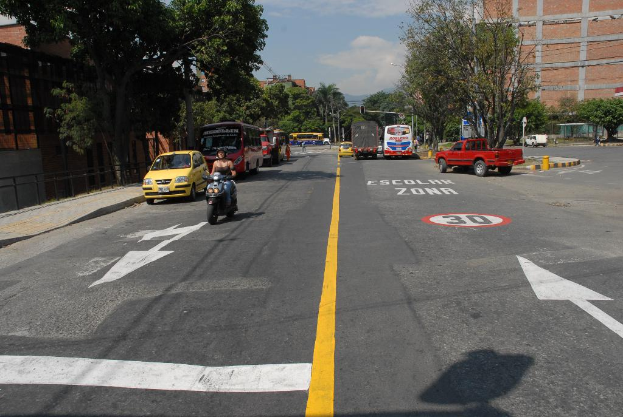 Eine Straßenszene mit Gebäuden, Bäumen, fahrenden Fahrzeugen, Schildern, Verkehrsampeln, einem blauen bewölkten Himmel und Warnschildern auf der Straße.