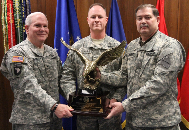 Drei Männer in Uniformen halten Auszeichnungen, mit Fahnen im Hintergrund und farbigem Text in der oberen linken Ecke.