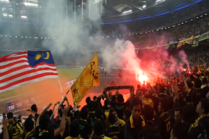 Eine große Menge in einem Stadion mit Fackeln und Rauch, einige halten Fahnen und eine Kamera, mit Schildern, die "Malaysia vs Malaysia Live Streaming" anzeigen.