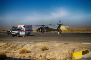 Ein Hubschrauber auf einem grasbewachsenen Straßenrand neben einem Rettungswagen geparkt, mit Menschen drumherum und Bergen und einem klaren blauen Himmel im Hintergrund.