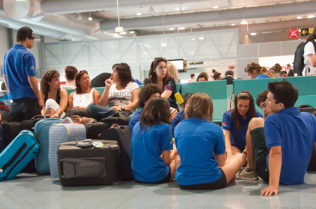 Ein belebter Flughafeninnenraum mit vielen Menschen, die auf ihre Flüge warten, Gepäck auf der linken Seite und Glastüren mit Richtungsschildern im Hintergrund.