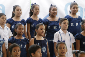 Gruppe von Kindern vor einem Mikrofon mit einem Banner im Hintergrund, die scheinbar als Teil eines Schulchors singen.