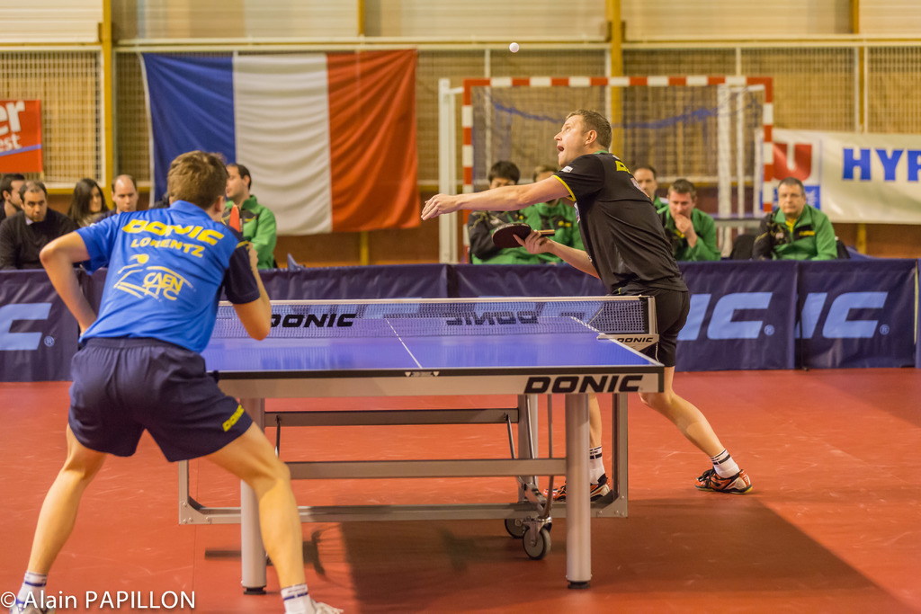 Zwei Personen stehen und halten Tischtennis-Schläger, während zwei andere in der Nähe sitzen; eine Flagge und Banner sind an der Wand dahinter sichtbar.