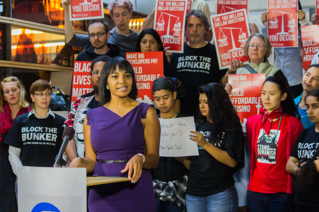 Eine Black Lives Matter-Aktivistin spricht vor einem Publikum mit Schildern und einem Mikrofon, während sie von Scheinwerfern und anderen Gegenständen im Hintergrund umgeben ist.