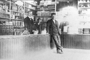 Drei Männer stehen an einem Tresen in einem Laden, umgeben von verschiedenen Gegenständen, mit einem Regal voller Bücher und anderen Gegenständen im Hintergrund, die scheinbar an einem Raub beteiligt sind.