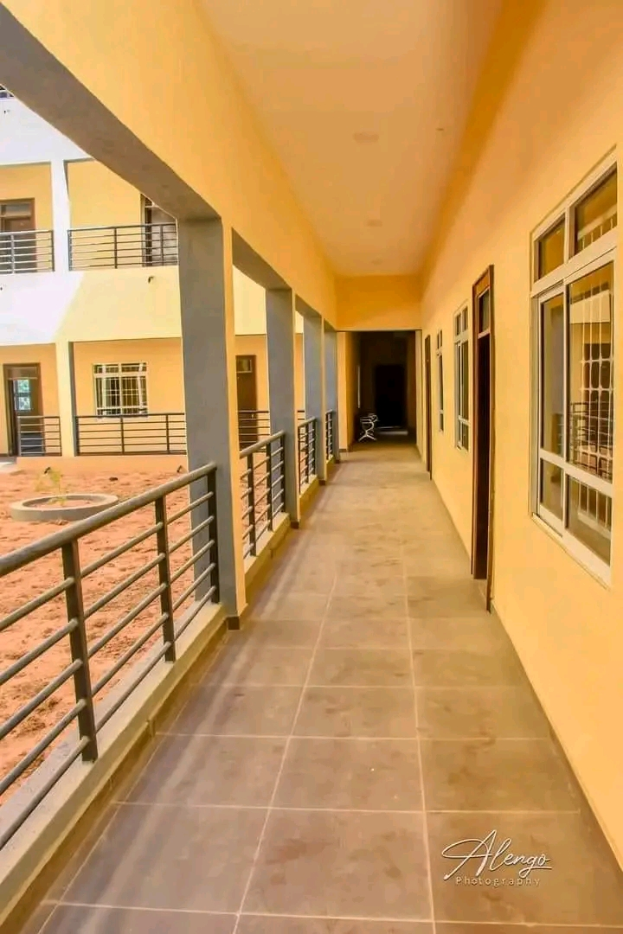 Ein Gebäude mit Fenstern, Türen, Säulen, Geländern, einem langen Flur mit einem gefliesten Boden und einem Balkon, sowie anderen Gebäuden und einem klaren blauen Himmel im Hintergrund und einem Text am unteren Rand, der "3-Zimmer-Wohnung zur Miete in Ntinda, Kampala" lautet.