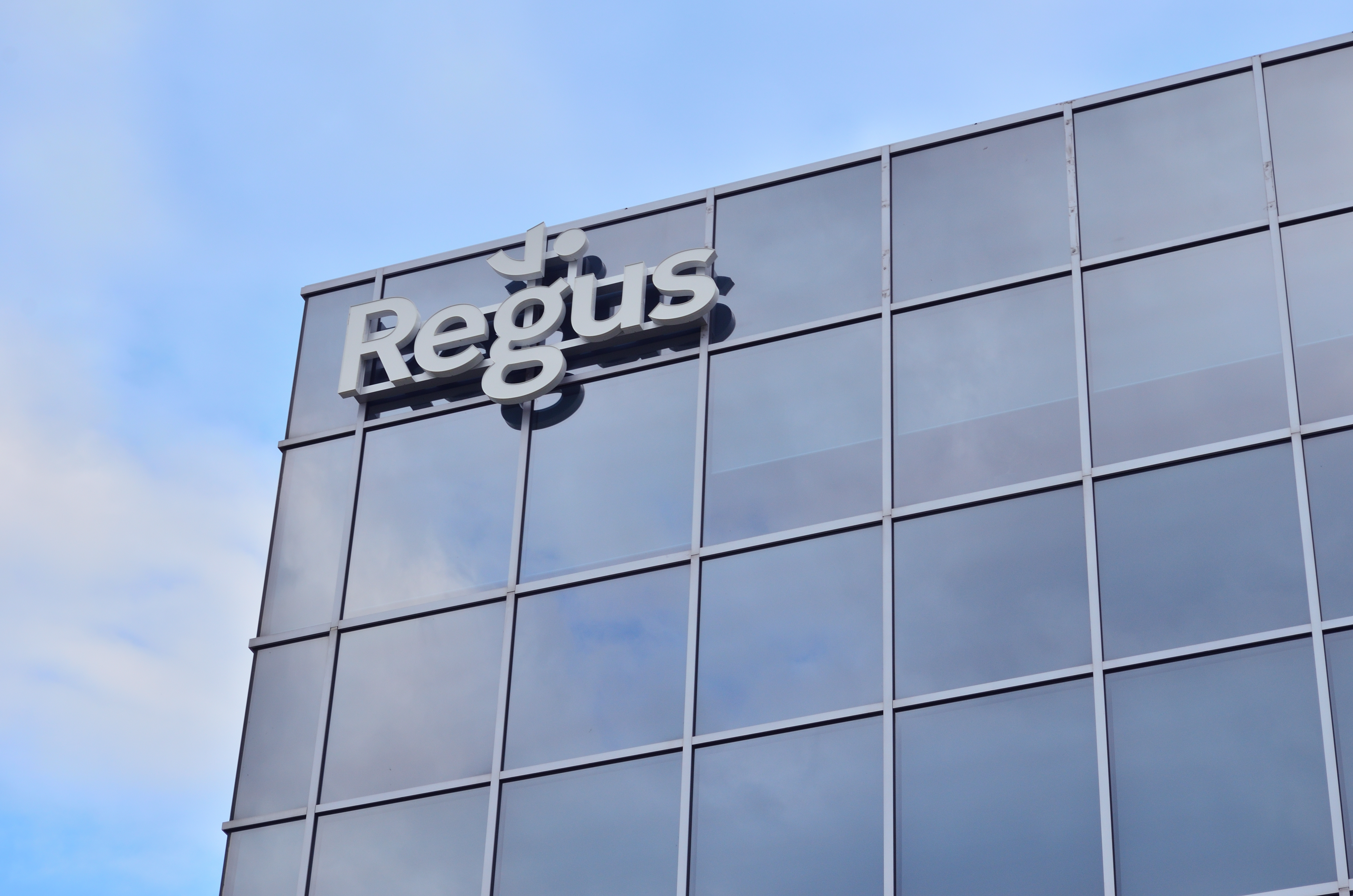 Ein Geb├Ąude mit einer Regus-Tafel an der Seite, umgeben von einem bew├Âlktem Himmel, mit dem Text "Regus investiert 1,5 Milliarden Dollar in US-Hauptstadt" darauf.