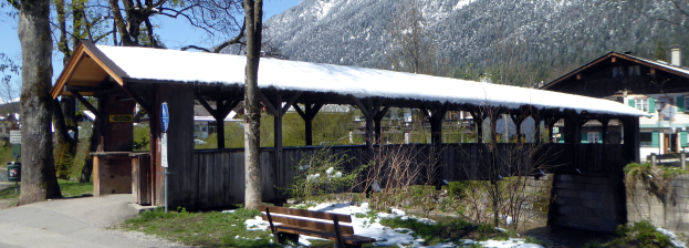 Ein hölzernes Unterschlupf mit schneebedecktem Dach, eine Bank davor, Pflanzen, Gras, eine Gruppe von Bäumen, Häuser mit Dächern und Fenstern, ein Schild, ein Müllcontainer, Pfosten, Hügel und ein bewölkter Himmel.