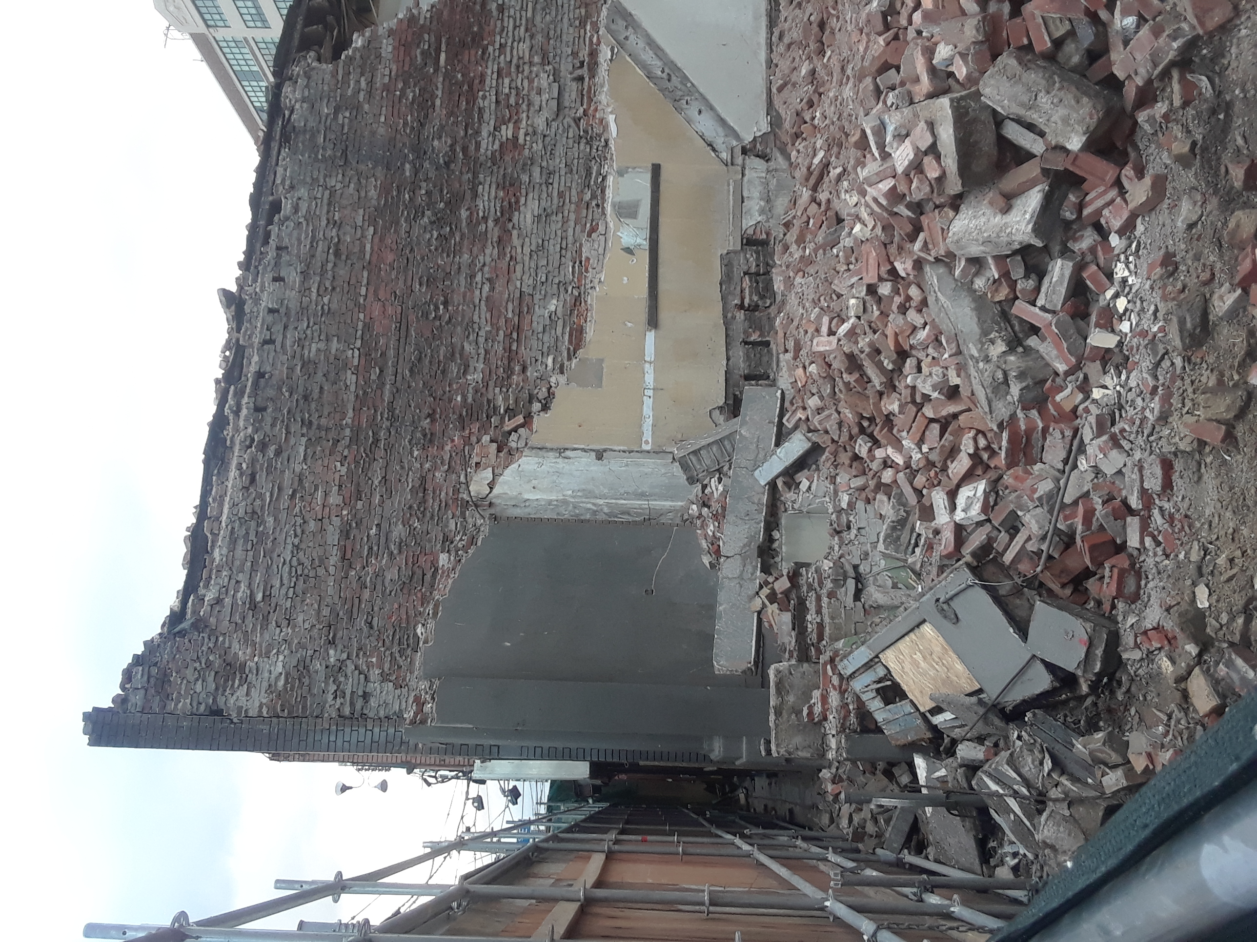 Teilweise abgerissenes Gebäude mit verstreuten Ziegeln und Metallstangen, mit anderen Gebäuden und einem klaren blauen Himmel im Hintergrund.