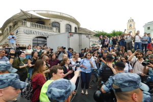 Große Gruppe von Menschen protestiert vor einem Gebäude, einige halten Kameras und Telefone, mit einem Fahrzeug, Bäumen und einem klaren blauen Himmel im Hintergrund.