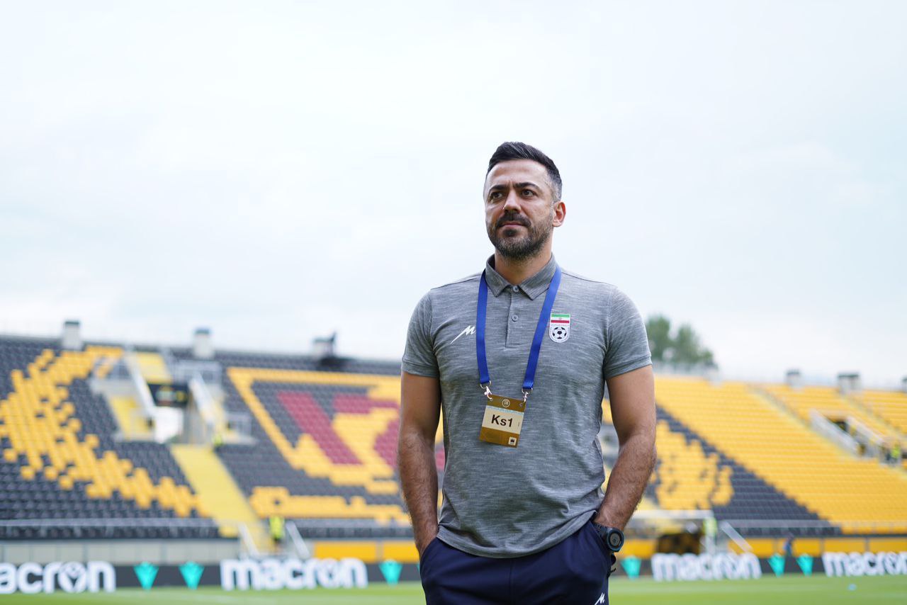 Ein als Ks1 identifizierter Mann, der Trainer der türkischen Nationalmannschaft, steht auf einem Fußballfeld in einem grauen T-Shirt und blauen Hosen. Im Hintergrund sind gelbe Stühle, ein Baum und ein klarer blauer Himmel zu sehen.