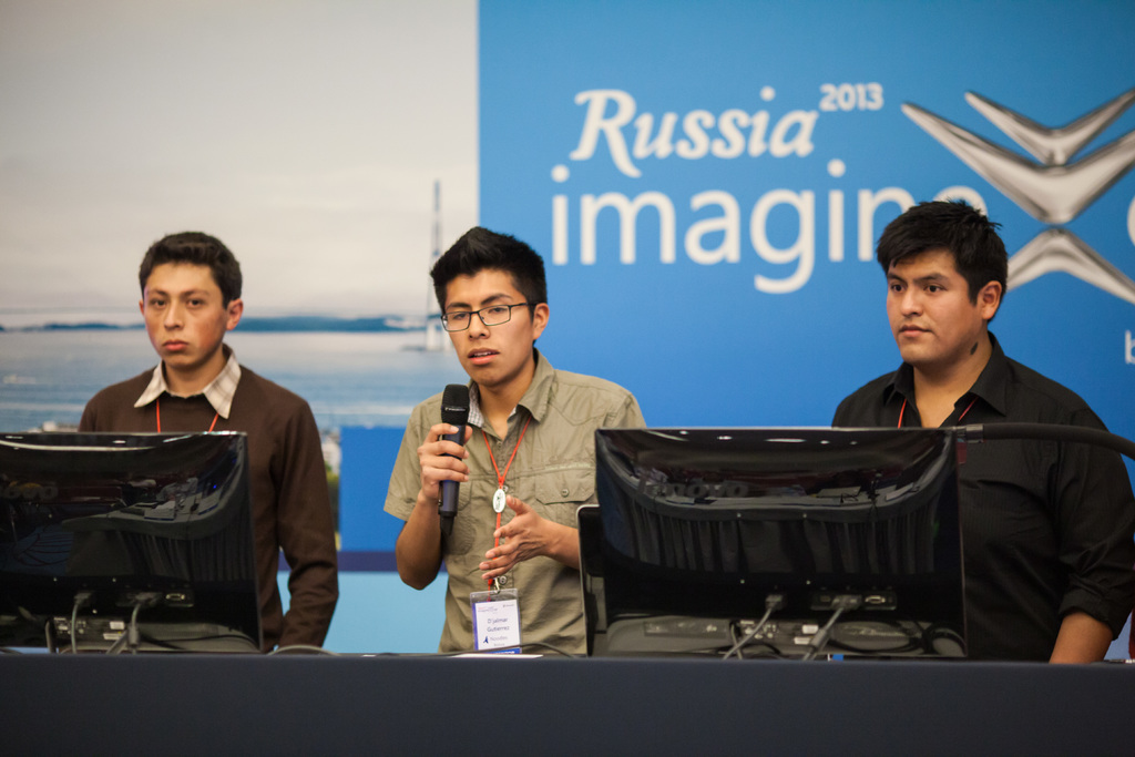 Drei Jungen stehen neben einem Schreibtisch mit zwei Computersystemen, einer spricht in ein Mikrofon und trägt eine ID-Karte mit roter Markierung, vor einer Wand mit einer Werbetafel, auf der "Russia Imagine 2013" steht.
