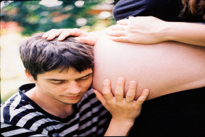 Ein Mann und eine Frau stehen nebeneinander, der Mann berührt sanft den Bauch der Frau, mit einem unscharfen Hintergrund, der das Paar und ihr ungeborenes Kind betont.