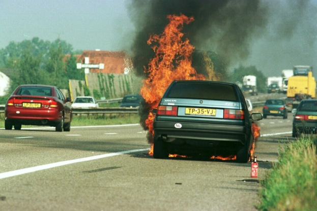 Ein Auto in Flammen neben der Straße mit anderen Fahrzeugen in der Nähe, Bäumen und Gebäuden im Hintergrund bei einem klaren blauen Himmel und einem Feuerlöscher auf dem Gras auf der rechten Seite.