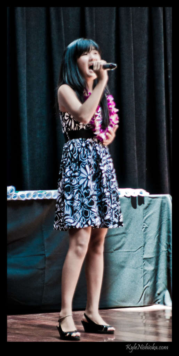 Eine Frau in einem bunten Kleid singt auf der B├╝hne in ein Mikrofon, mit einem tisch voller Papiere und einem Vorhang im Hintergrund.