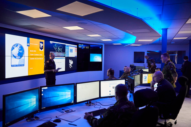 Eine Gruppe von Menschen an Schreibtischen mit mehreren Monitoren, Tastaturen und Papieren, während eine Person mit einem Papier in der Hand steht, in einem Raum mit wandmontierten Bildschirmen und Deckenleuchten, was auf eine Cyber-Sicherheitszentrale hinweist.