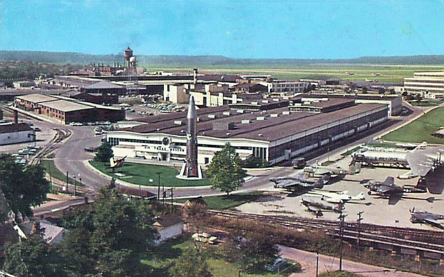 Altes Schwarz-Weiß-Foto eines Flughafens mit geparkten Flugzeugen, Gebäuden, Bäumen, Straßen, Fahrzeugen, Strommasten, Gras, Bahnschienen, Türmen, Hügeln und einem klaren Himmel mit Text unten.