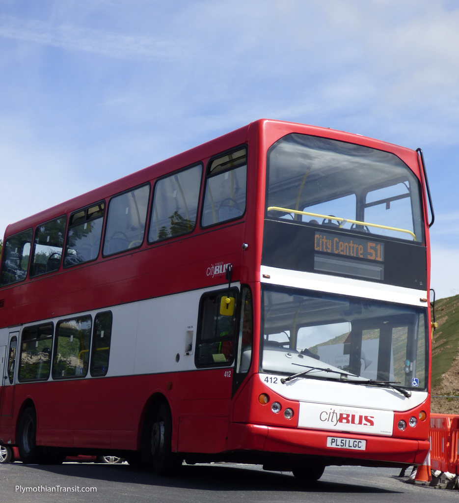 Ein roter Doppeldecker-Stadtbus ist auf der Straße unterwegs mit einem Verkehrskegel davor und einem Hügel im Hintergrund.