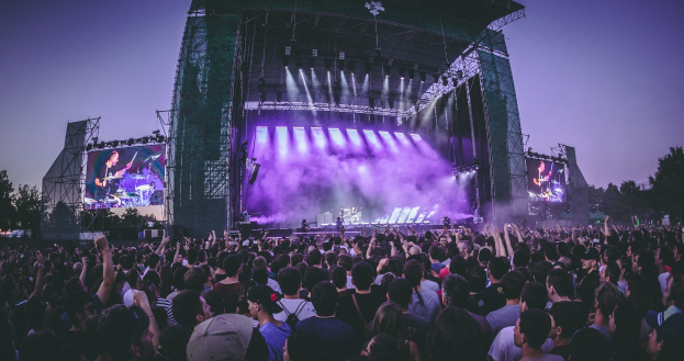 Eine große Menge bei einem Musikfestival schaut auf einer hell erleuchteten Bühne mit Instrumenten und Bildschirmen, die vor einem Hintergrund aus Bäumen, einem Gebäude und einem klaren blauen Himmel liegt.