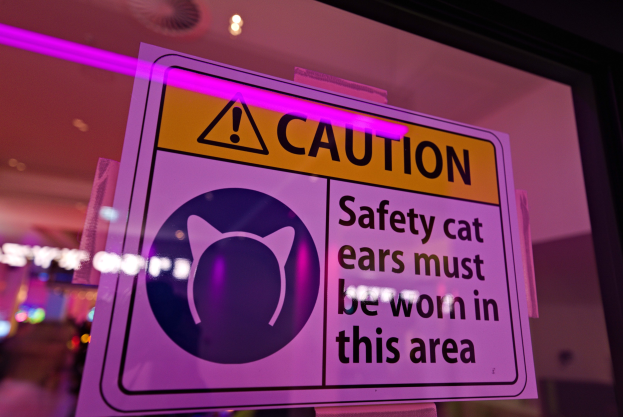Hellgelbes Warnschild in einem Ladenschaufenster mit der Aufschrift "Vorsicht: Sicherheits-Katzenohren müssen in diesem Bereich getragen werden" vor einem unscharfen Hintergrund.